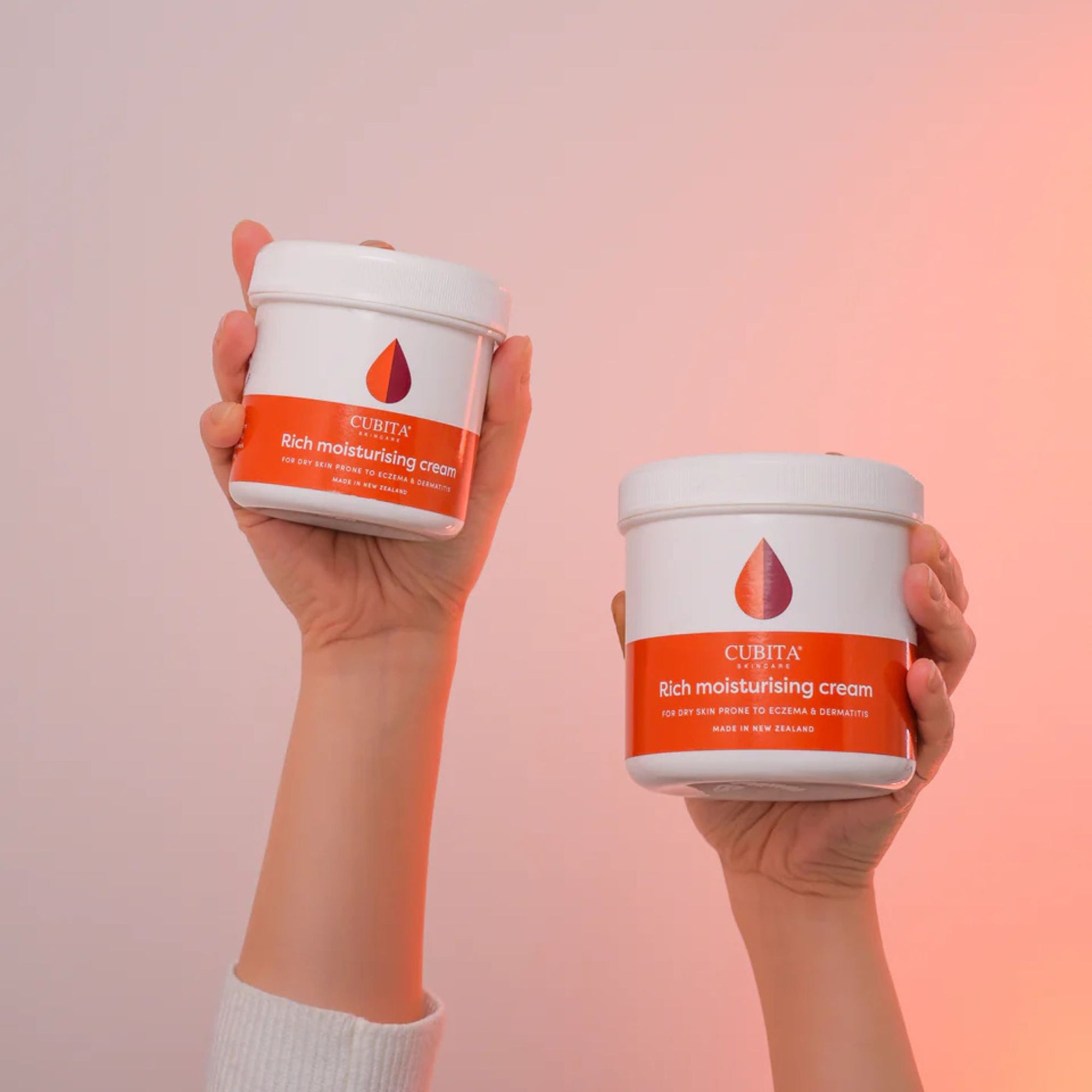 Two hands holding jars of Cubita rich moisturizing cream against a gradient background