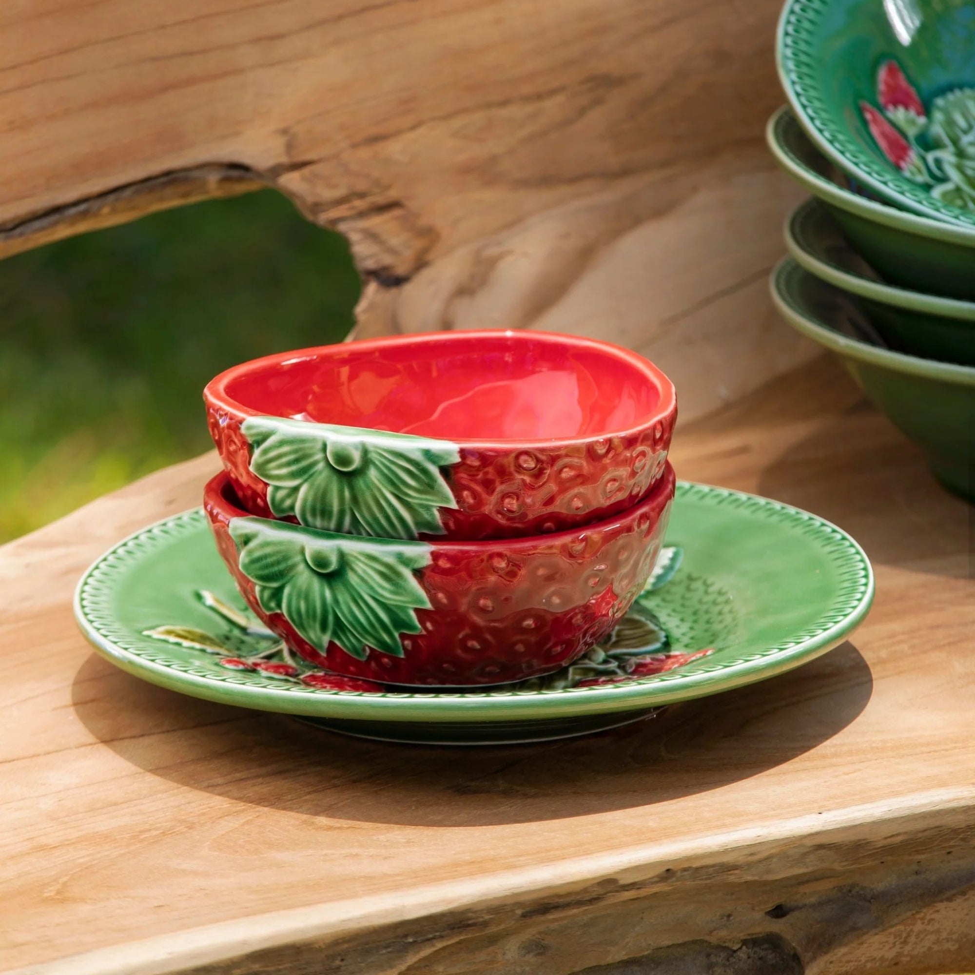 Stack of red ceramic bowls with green strawberry design on a matching green plate, placed on a wooden surface.