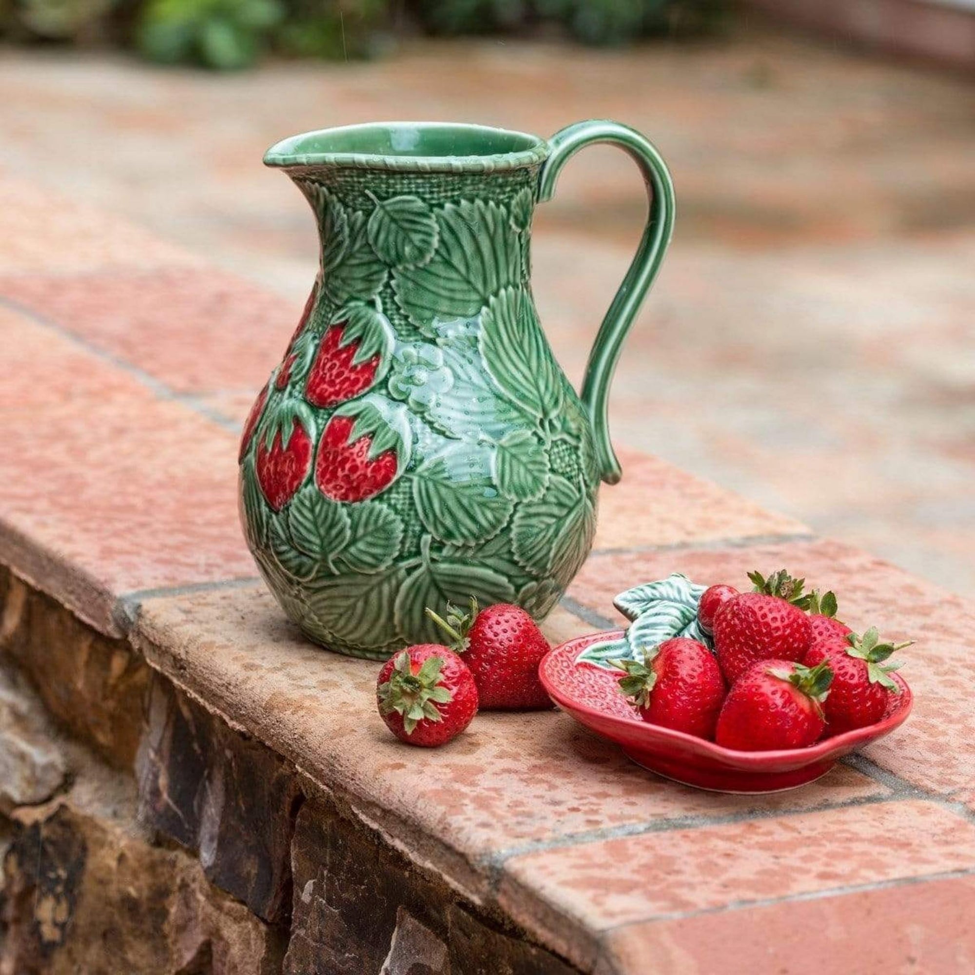 Bordallo Strawberries Pitcher on a stone surface with strawberries around it.